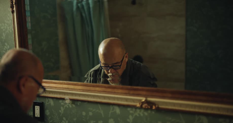 Man with glasses shaves with electric razor in front of mirror in bathroom with floral wallpaper