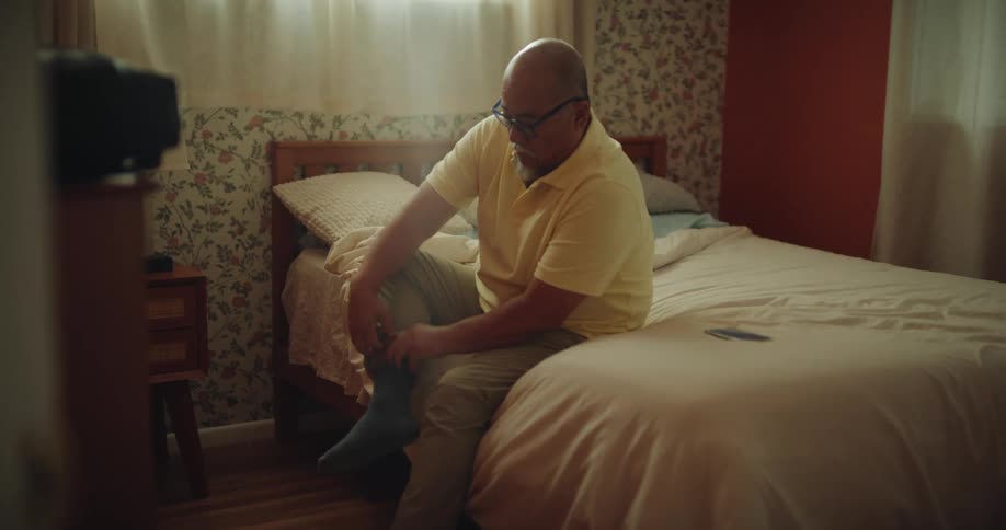 Man finishes getting dressed in his bedroom by putting on socks then gathers his things to leave
