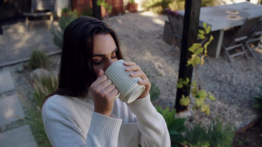 A young woman outdoors in a backyard at midday sipping a cup of coffee in the warm daylight