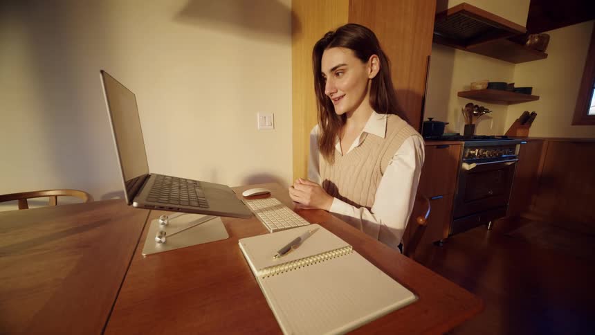 Young adult woman sitting down on a chair talking in front of a laptop computer on a wooden desk indoors during dusk or dawn