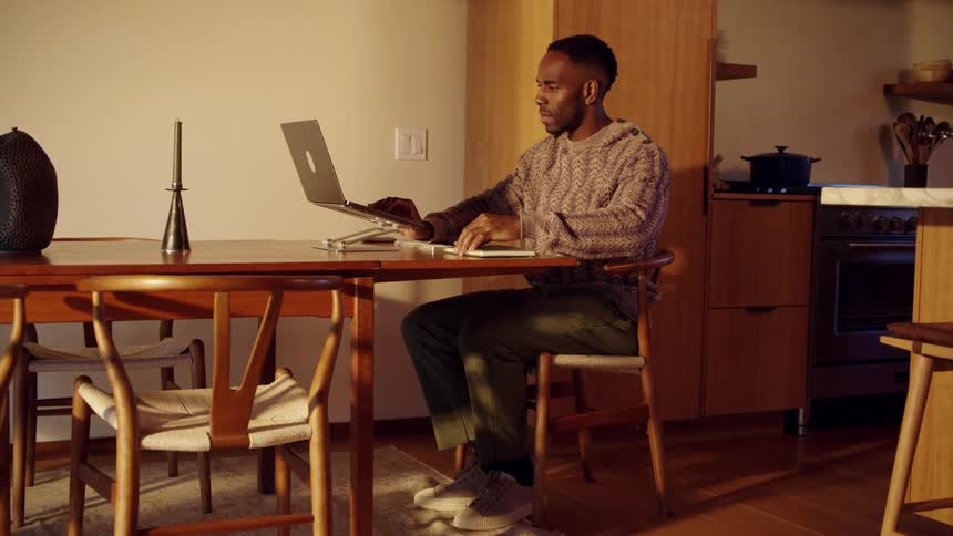 Man sits at a table rubbing his hands together then opens the lid of his laptop computer and writes in a notebook