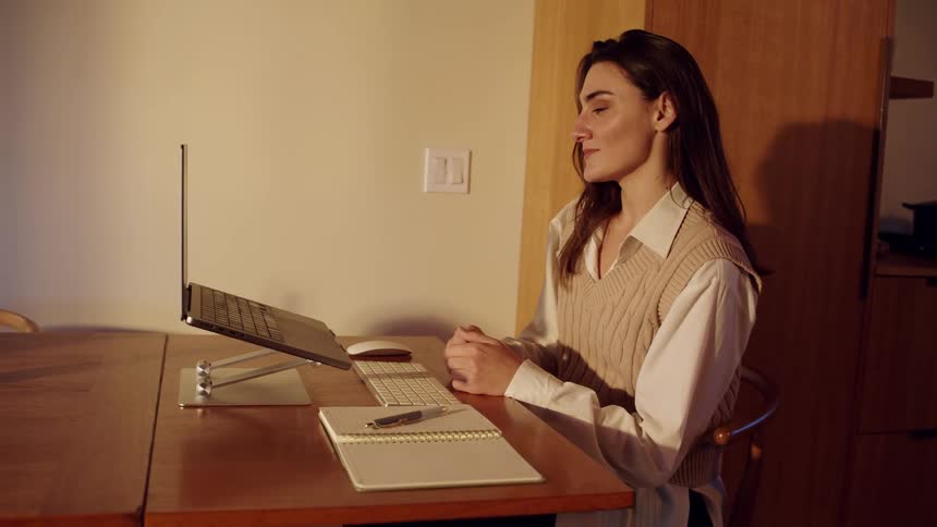 Woman in beige vest and white shirt works focused on computer at wooden desk in cozy setting