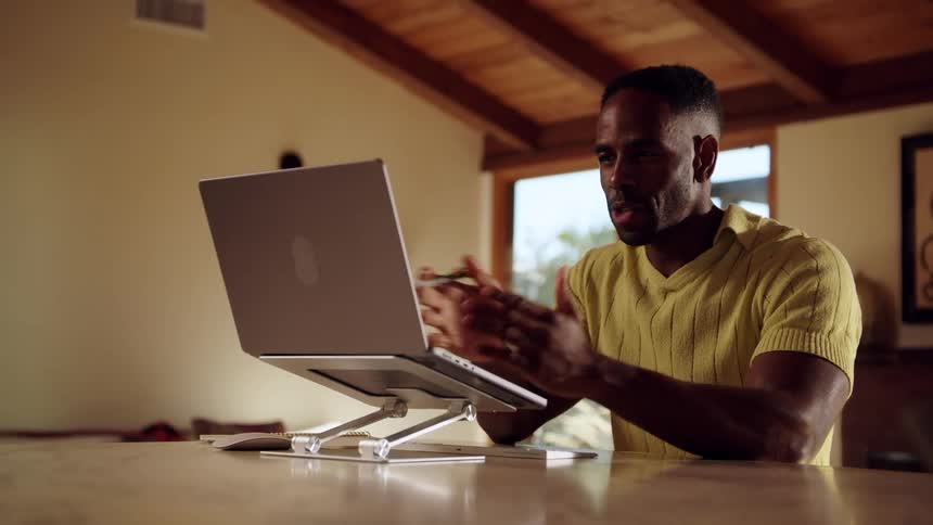 Person in yellow shirt gestures while working on elevated laptop in sunlit room with wooden beams