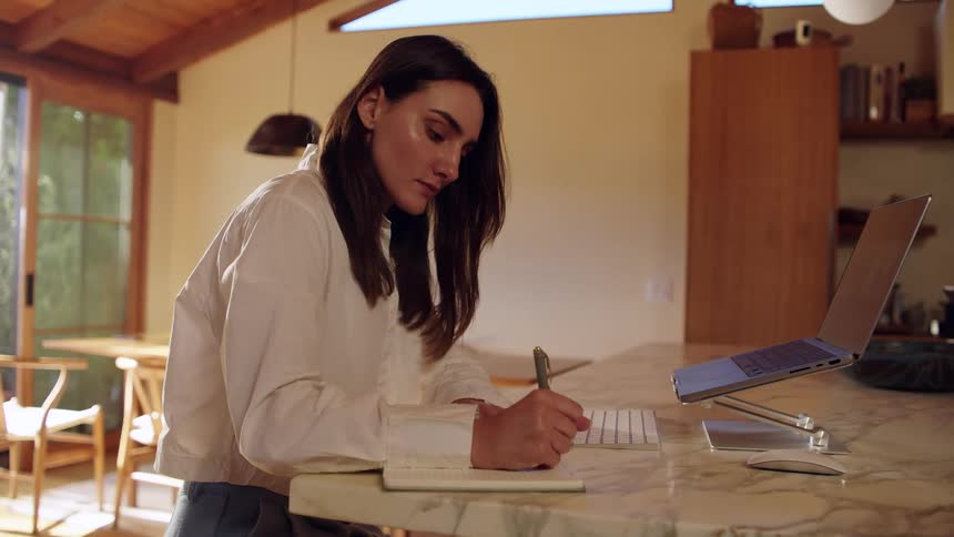 Woman sits at a table with a laptop computer typing on a keyboard and stops to write on paper