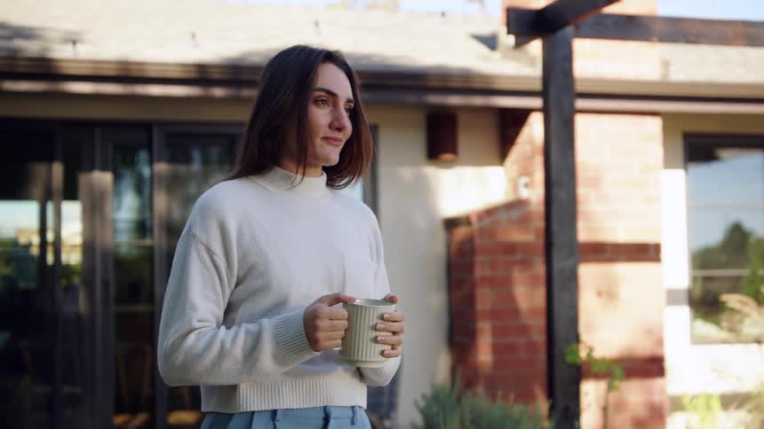 Woman drinks a beverage from a mug while standing outdoors near a house