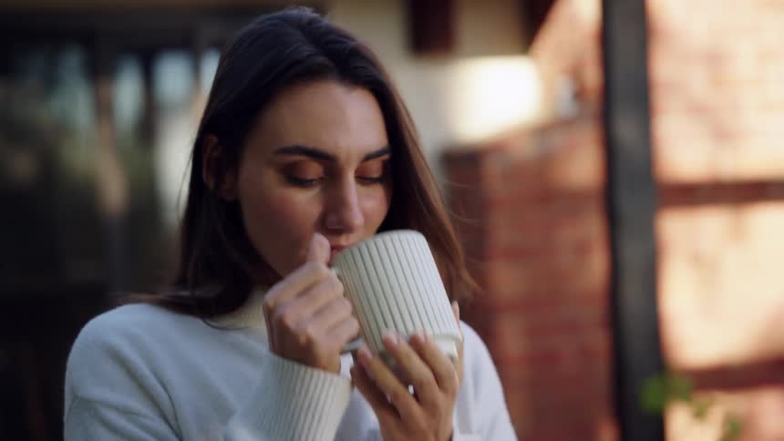 Woman drinks a hot beverage from a mug in an outdoor setting near a building