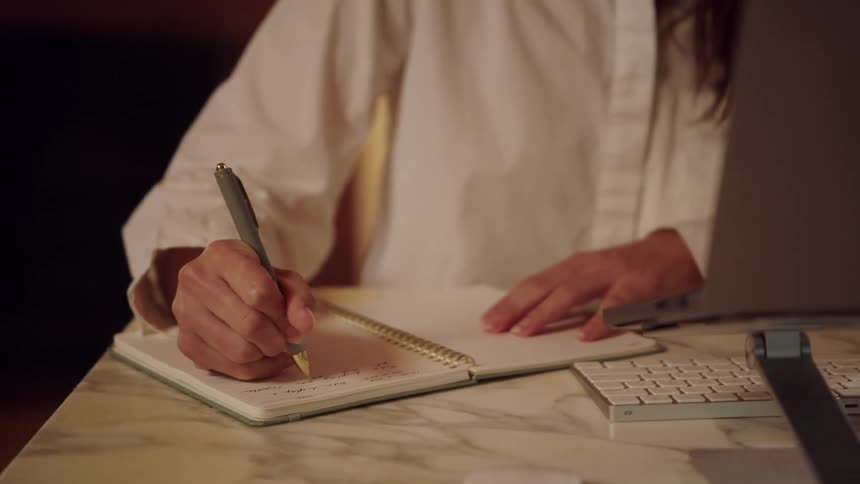 Young adult woman writing with a pen on a notebook while talking in front of a laptop computer over a desk indoors during day time