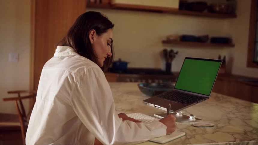 A woman in a white shirt sits at a marble kitchen island writing in a notebook while looking at a laptop with a green screen mounted on a silver riser in a warm sunlit home interior