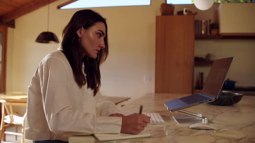 A woman writes and types on a laptop at a kitchen counter