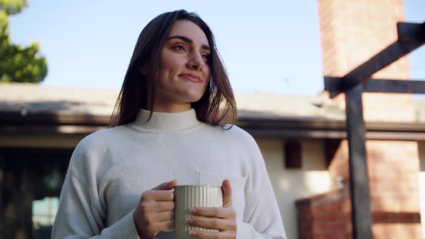 Woman holds a mug with both hands outdoors near a residence