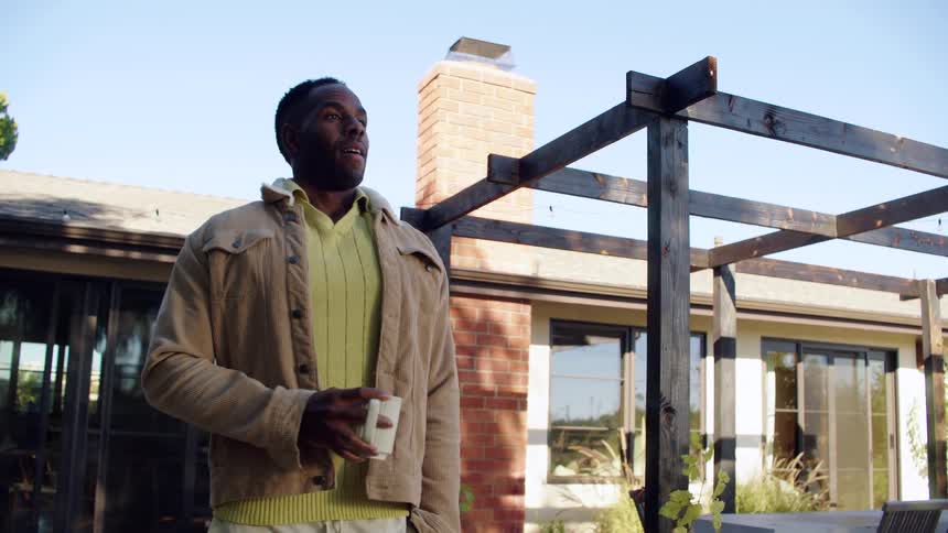 A man in a tan jacket and yellow polo shirt walks through a sunny backyard garden and takes a sip from a white mug