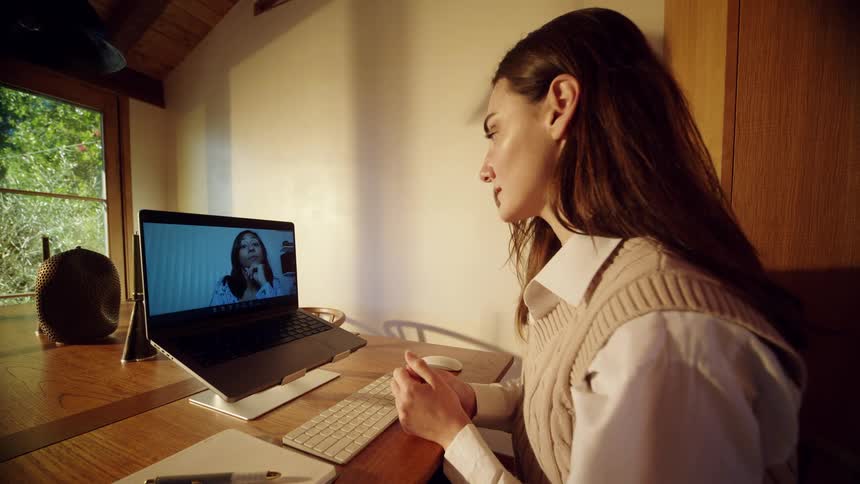A woman with long dark hair wearing a white shirt and tan vest participates in a video call on a laptop while sitting at a wooden desk in a sunlit room