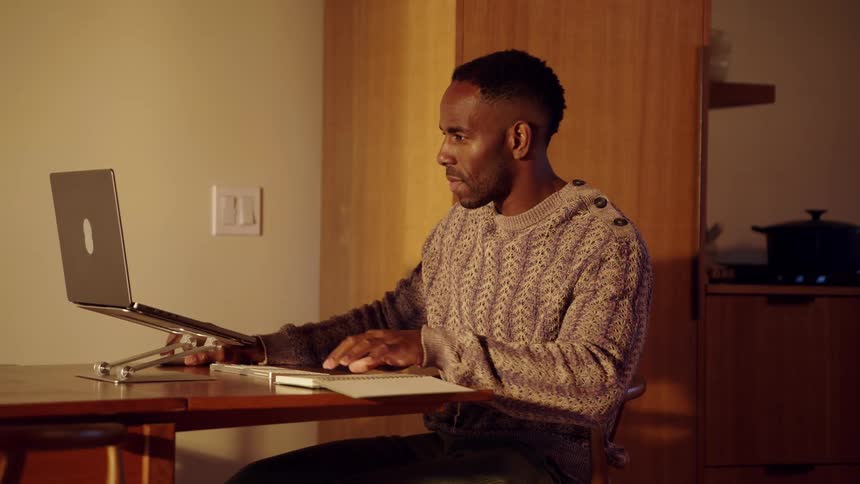 Man sitting at a table holds his notebook as he opens the lid of his laptop computer and writes in the notebook