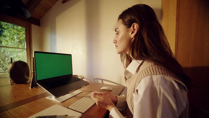 A young woman in a beige vest sits at a wooden desk gesturing and talking while looking at a laptop screen in a warm sunlit home office