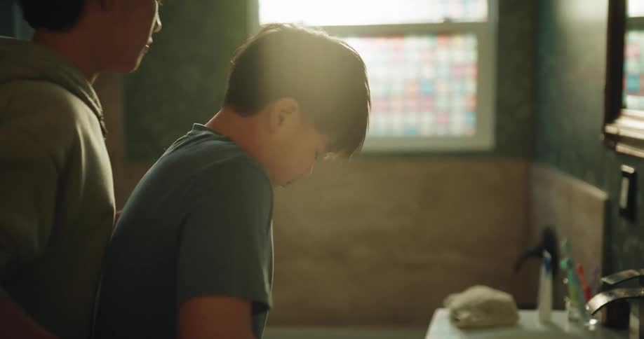 In a sunlit bathroom a young boy struggles to open a container of deodorant until an older boy enters to