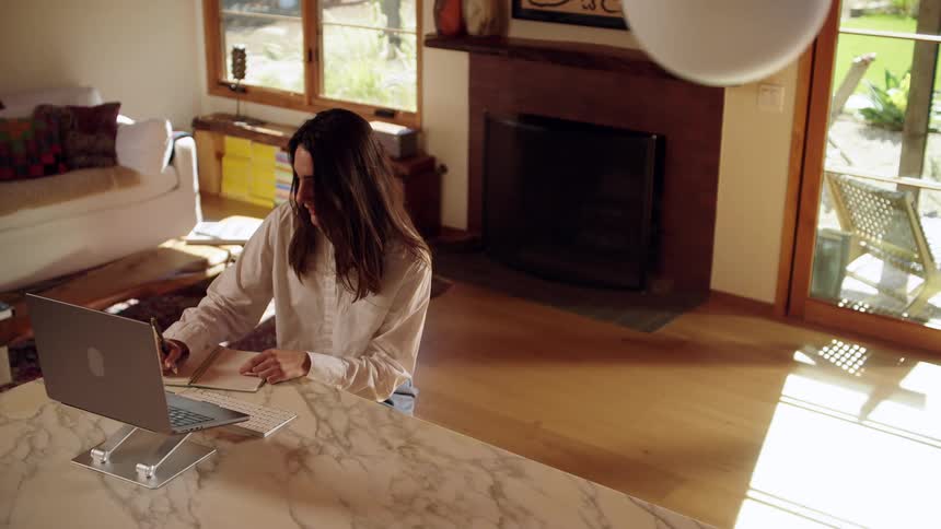 A young woman sitting in the dining area of her living room at midday taking notes during a conference call on her laptop while gesturing with her arm as she speaks