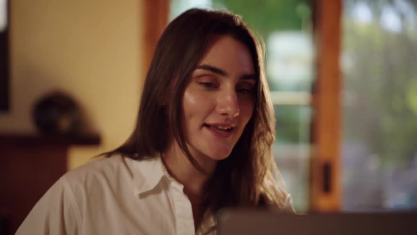 A young woman sitting in her living room at midday facing her computer as she participates in an interview