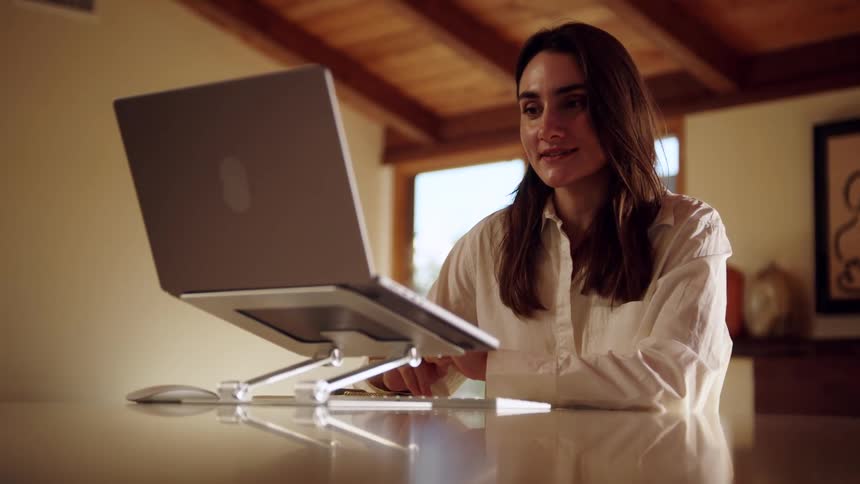 A woman sits at a wooden table in a warmly lit room engaging in a video call on her laptop she gestures with her hands while speaking and smiling with her computer elevated on a silver adjustable stand