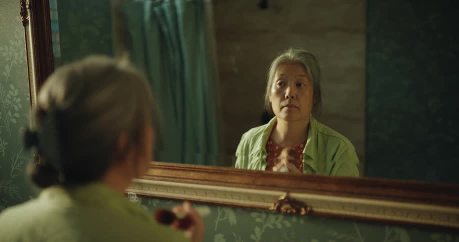 An elderly asian woman stands before a bathroom mirror meticulously applying face powder with a large brush before putting on her glasses and adjusting her green outer shirt