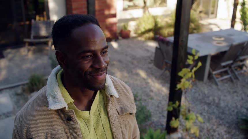 Young adult black man standing on a lawn next to a house opening and closing his eyes with a nervous facial expression and then smiling outdoors during day time