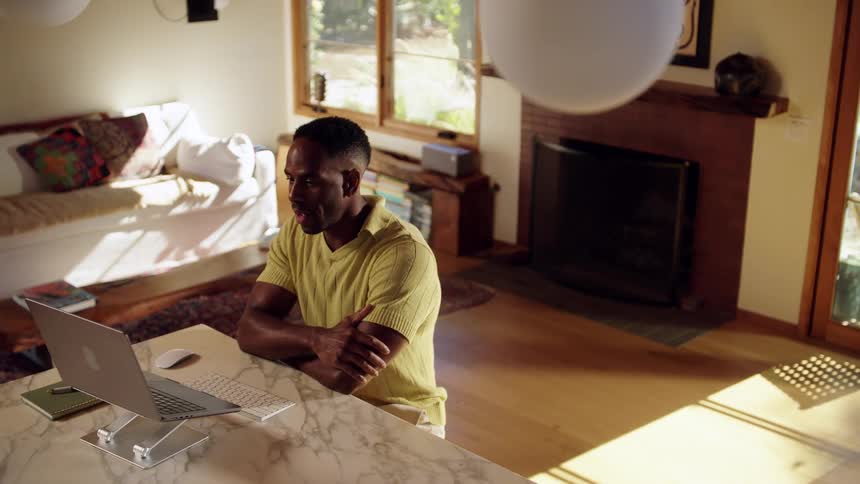 A young man sitting at a desk in his living room at midday speaking to an interviewer on a conference call with his laptop open in front of him