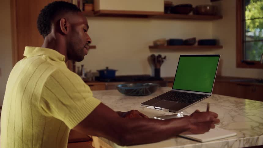 A man wearing a yellow shirt sits at a marble kitchen counter and participates in a video call on a laptop with a green screen while writing in a notebook