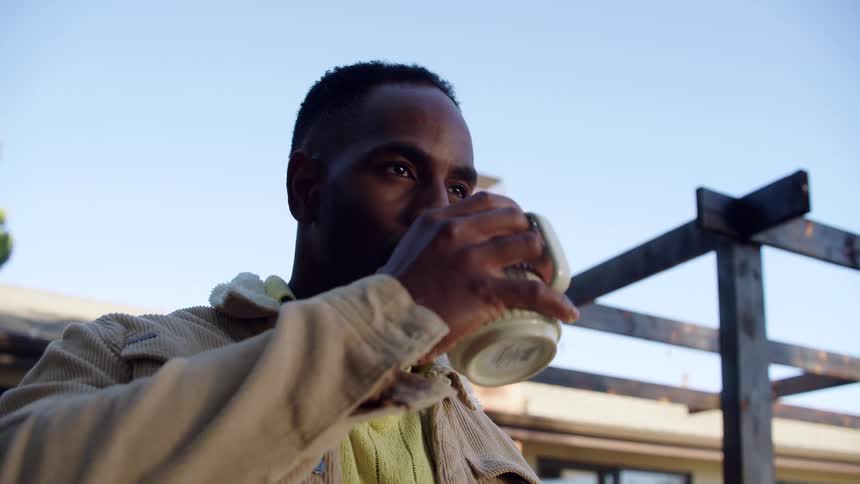 A smiling man wearing a tan corduroy jacket over a yellow sweater stands outside under a clear sky enjoying a drink from a ceramic mug while looking thoughtfully into the distance near a wooden pergola