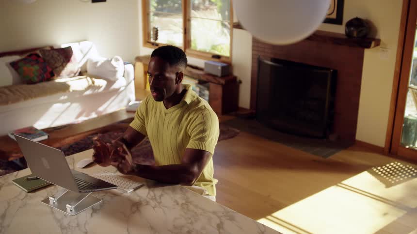A man wearing a yellow polo shirt sits at a marble kitchen counter and gestures with his hands while participating in a video call on a laptop in a sunlit living room