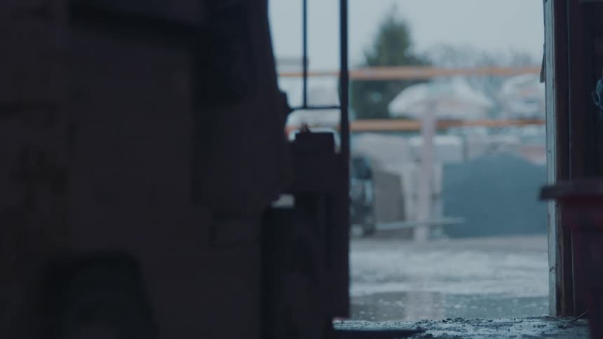 A worker in a blue shirt pulls down a large white sectional garage door to close a warehouse or workshop while heavy machinery and rain are visible outside