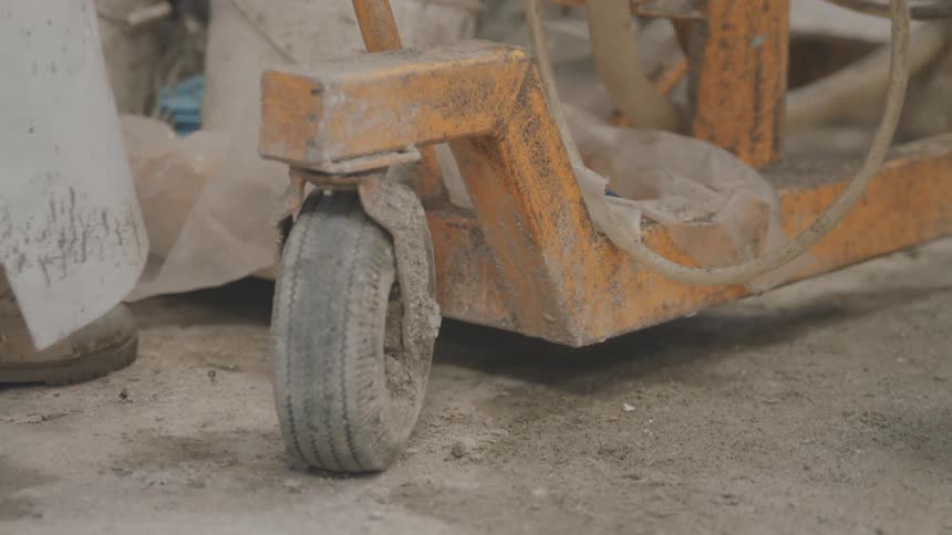 A close view shows the dusty boots of a worker pulling a heavy orange industrial machine on a concrete floor in a factory