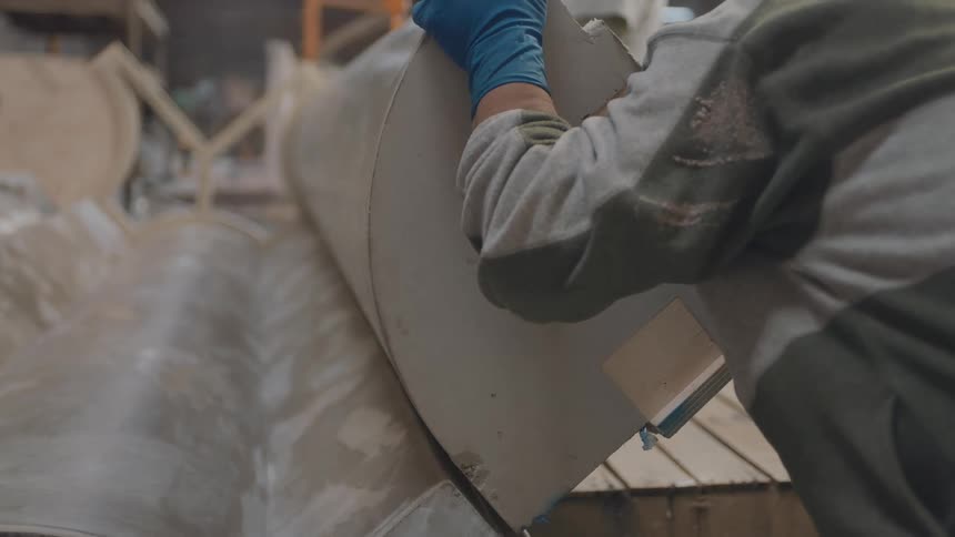A worker wearing blue gloves and a grey baseball cap carefully aligns a large cylindrical metal component in an industrial manufacturing facility