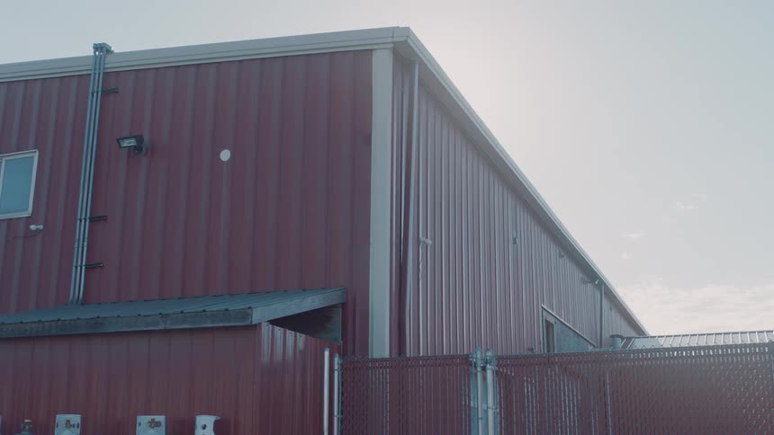 A red industrial building with corrugated metal siding stands under a bright sky next to a tall chain link fence