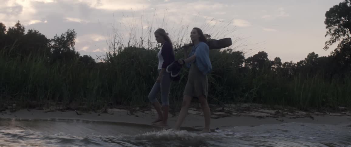 Two women walk barefoot on a lakeshore