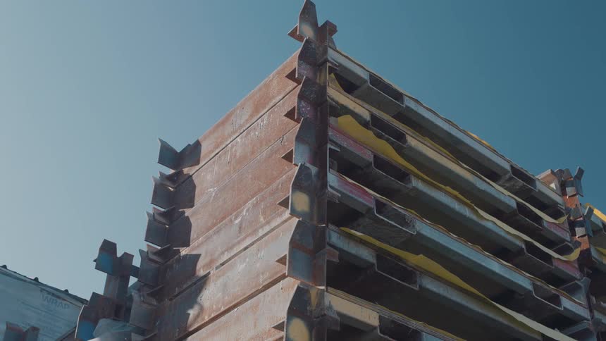 A stack of rusty steel industrial beams with sharp edges sits outdoors under a clear blue sky on a bright day