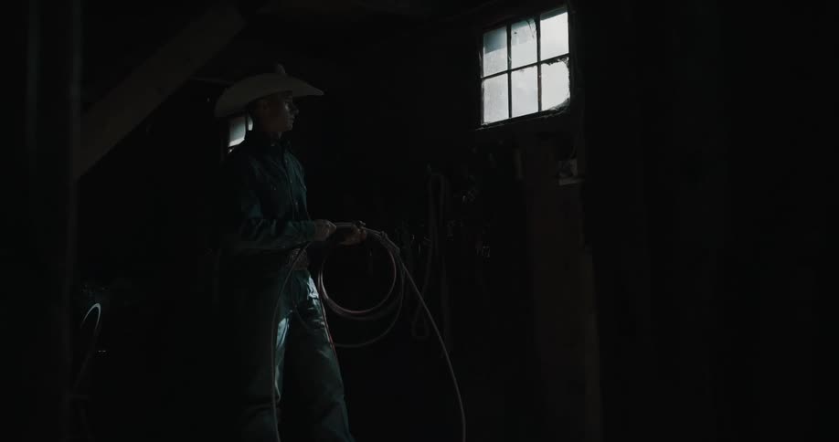 Person holds a lasso inside a dim stable lit by a window