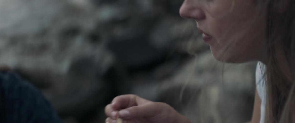 A woman rolls a cigarette on a rocky beach