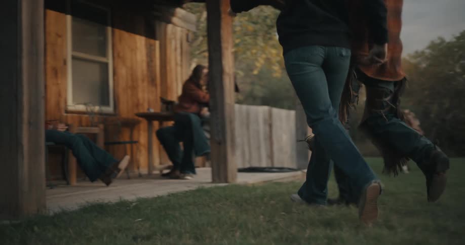 Young adult man and woman running through a courtyard surrounded by farm horses in a rural area towards a porch where other people are sitting down on chairs outdoors during dusk or dawn