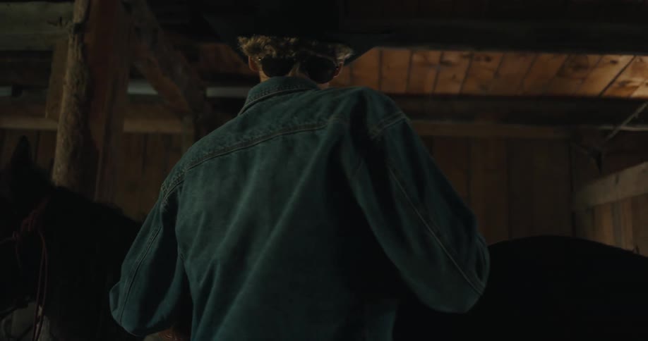 Young adult man wearing a cowboy hat and a denim jacket inspecting the saddle on a horse on a stable indoors during day time