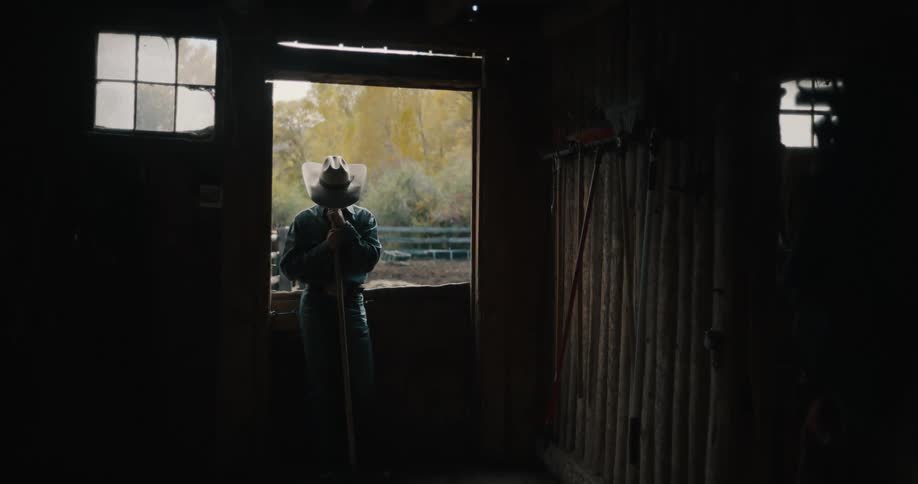 A man wearing a widebrimmed hat and denim stands silhouetted in a barn doorway while leaning on a pitchfork looking out toward a sunlit autumn landscape from the shadows of a rustic wooden stable filled with hanging tools