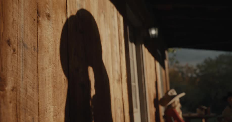 Shadows of a man and a woman wearing cowboy hats are cast onto the side of a wooden building as they lean in to share a kiss in the warm afternoon light