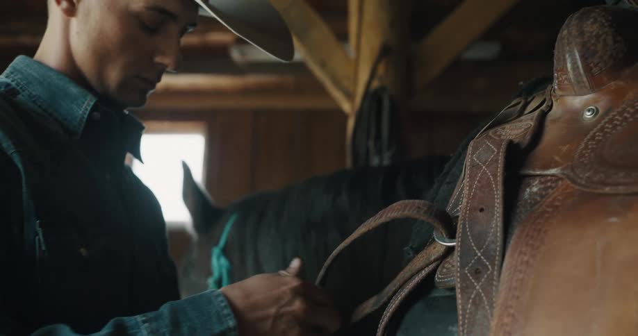 Person adjusts a leather saddle on a horse inside a wooden stable