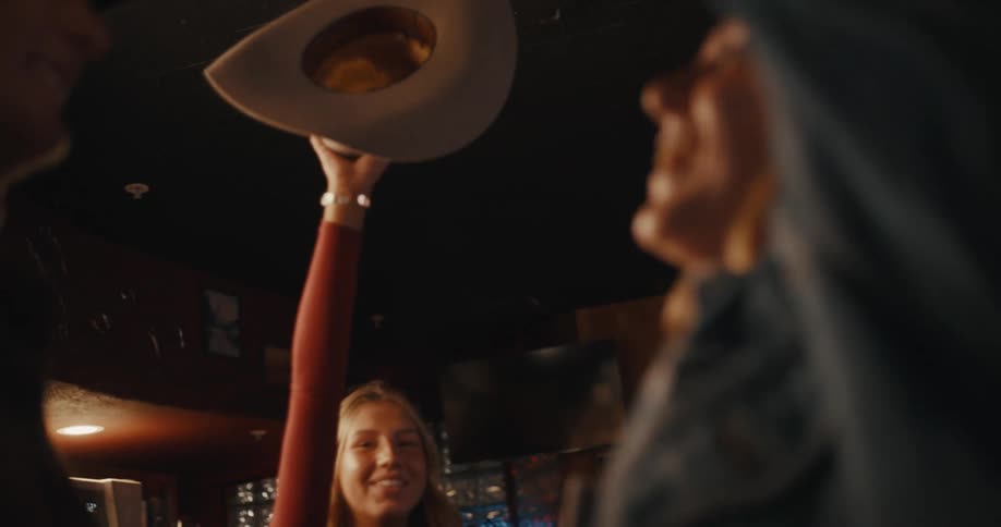A man and a woman wearing cowboy hats laugh and dance together in a crowded bar before leaning in close to each other under warm interior lighting
