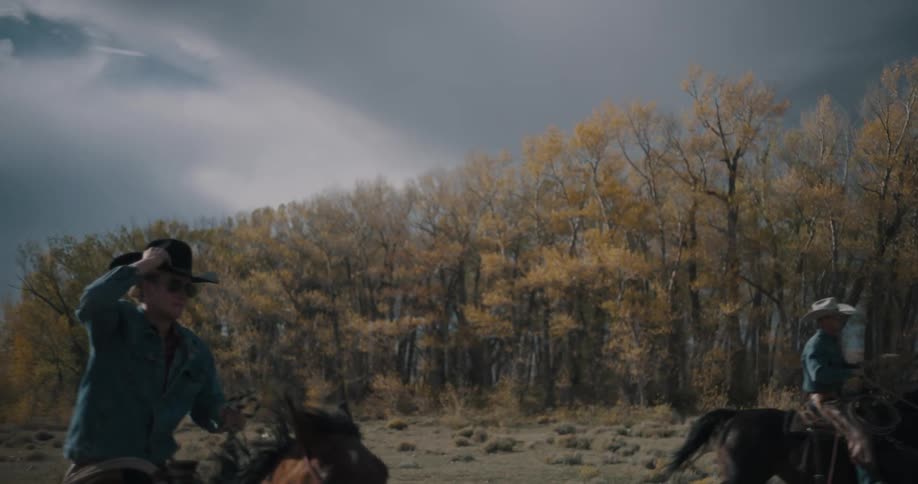Riders ride horses in an open area while one of them raises a hat in front of autumn trees