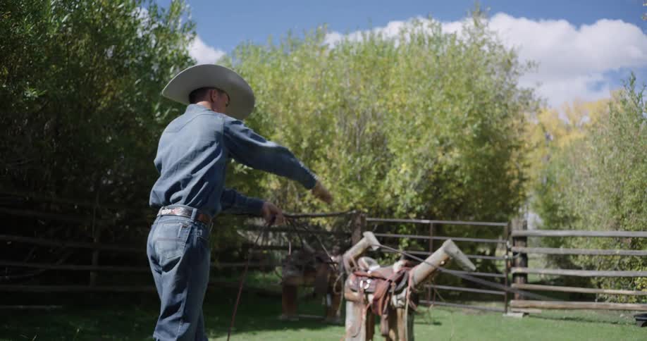 A cowboy in a denim outfit and hat practices throwing a lasso towards a wooden roping dummy in a grassy fenced in area