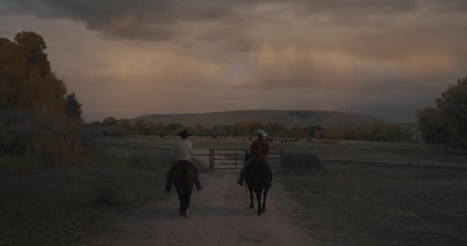 Two adult men riding horses on a path next to a wooden fance in a ranch with hills and trees on the background outdoors during dusk or dawn