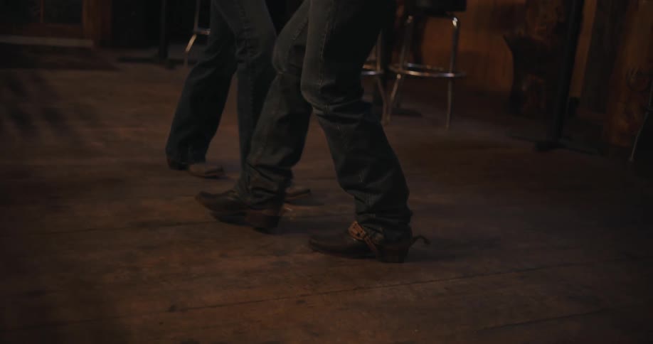 In a dimly lit bar two people in boots and jeans line dance on a wooden floor