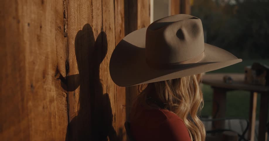 A woman with blonde hair wearing a beige cowboy hat sits in profile against a sunlit wooden wall