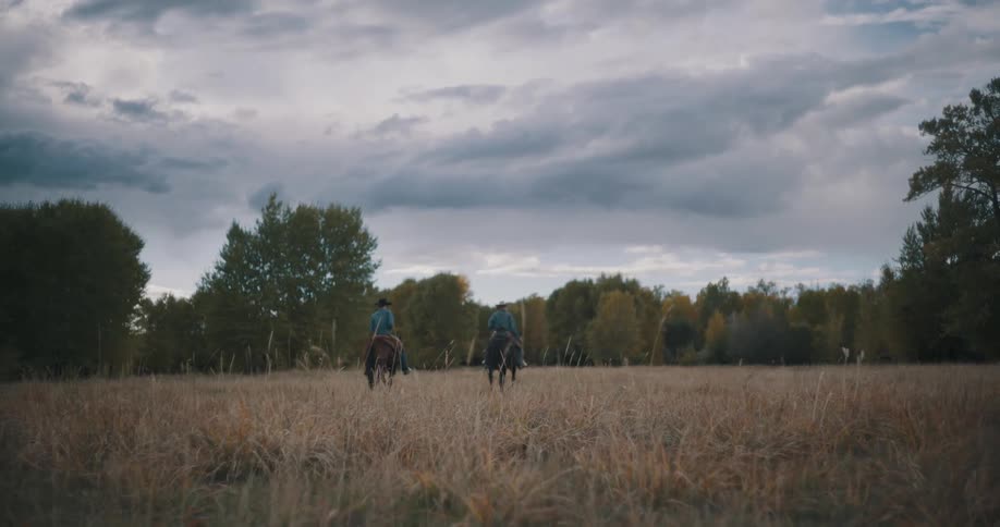 Riders travel on horseback across an open field toward trees under a cloudy sky