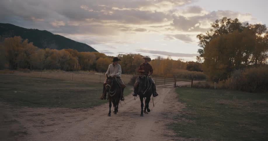 Two men ride horses on a rural dirt road with trees in fall colors and mountains in the distance