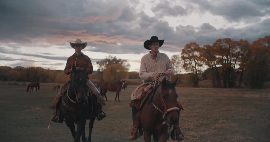 Two riders ride horses across an open field at dusk with trees in the background